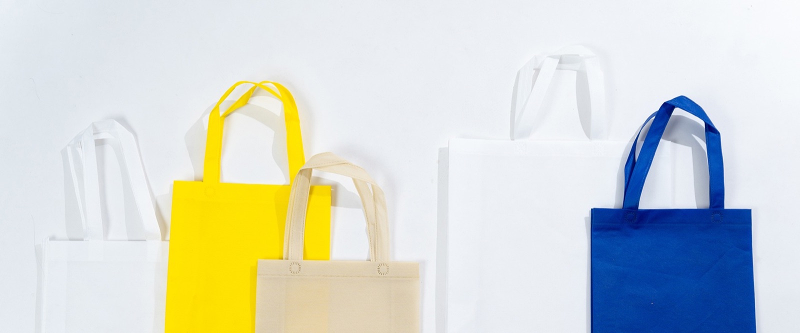 Five plain non woven tote bags in white, yellow, cream and blue, standing upright